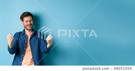 Image of happy man celebrating success, shouting yes and shaking clenched fists, achieve goal and triumphing, winning and dancing, standing on blue background Image of happy man celebrating success, shouting yes and shaking clenched fists, achieve goal and triumphing, winning and dancing, standing on blue background 99051934