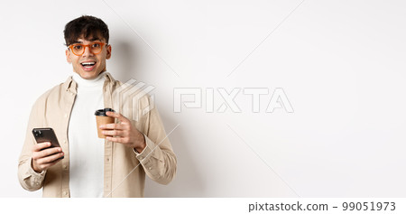 Cheerful smiling young man in glasses drinking coffee and holding smartphone, looking excited at camera, standing on white background Cheerful smiling young man in glasses drinking coffee and holding smartphone, looking excited at camera, standing on white background 99051973