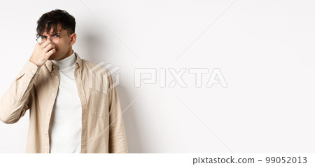 Image of disgusted guy shut his nose from awful smell, looking at something disgusting and stinky, standing on white background Image of disgusted guy shut his nose from awful smell, looking at something disgusting and stinky, standing on white background 99052013