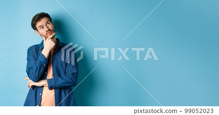 Pensive young man touching face and looking aside at empty space, imaging something, thinking off plan, standing on blue background Pensive young man touching face and looking aside at empty space, imaging something, thinking off plan, standing on blue background 99052023