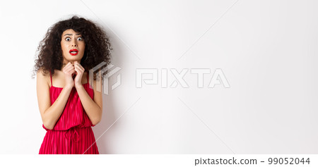 Scared caucasian woman trembling from fear, wearing red dress and staring anxious at camera, standing over white background 99052044
