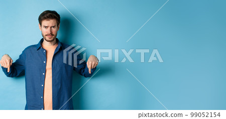Concerned young man, frowning and looking worried, pointing fingers down, showing something bad, standing on blue background Concerned young man, frowning and looking worried, pointing fingers down, showing something bad, standing on blue background 99052154