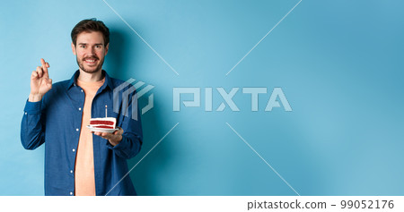 Celebration and holiday concept. Hopeful young man making wish on birthday cake, cross fingers for good luck and smiling positive, standing on blue background Celebration and holiday concept. Hopeful young man making wish on birthday cake, cross fingers for good luck and smiling positive, standing on blue background 99052176