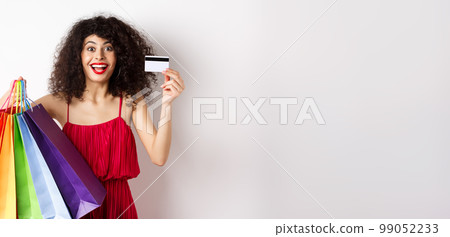 Stylish caucasian woman with curly hair and red dress, showing shopping bags and her plastic credit card, smiling amused, standing over white background Stylish caucasian woman with curly hair and red dress, showing shopping bags and her plastic credit card, smiling amused, standing over white background 99052233