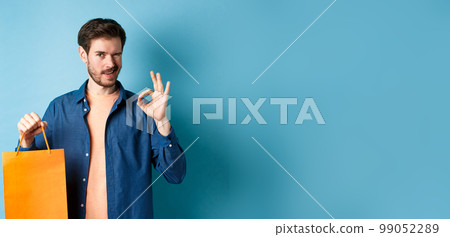 Modern man in casual outfit showing shopping bag and okay sign, winking at camera, recommending shop, standing on blue background Modern man in casual outfit showing shopping bag and okay sign, winking at camera, recommending shop, standing on blue background 99052289