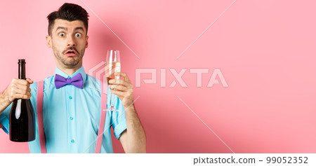 Holidays and celebration concept. Confused man staring startled at camera while pouring glass of champagne, standing puzzled over pink background Holidays and celebration concept. Confused man staring startled at camera while pouring glass of champagne, standing puzzled over pink background 99052352
