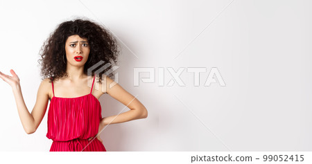 Upset pretty woman with curly hair and red dress, raising hand and look confused, frowning disappointed, talk about unfair things, standing against white background 99052415