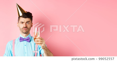 Holidays and celebration concept. Troubled young man in party hat, frowning with doubtful face, raising glass of champagne perplexed, standing on pink background 99052557