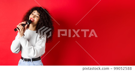 Leisure and hobbies concept. Stylish young woman singing karaoke, looking aside and holding microphone, performing song, standing on red background 99052588