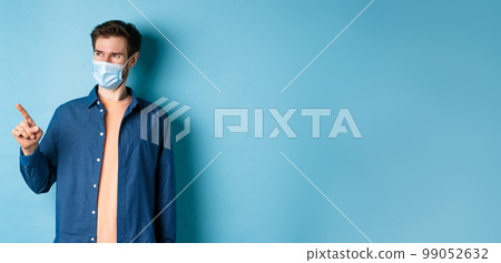 Handsome young man wearing medical mask from coronavirus, pointing finger left and looking satisfied, smiling at empty space, blue background Handsome young man wearing medical mask from coronavirus, pointing finger left and looking satisfied, smiling at empty space, blue background 99052632