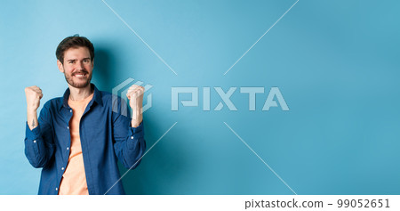 Image of happy man celebrating success, shouting yes and shaking clenched fists, achieve goal and triumphing, winning and dancing, standing on blue background Image of happy man celebrating success, shouting yes and shaking clenched fists, achieve goal and triumphing, winning and dancing, standing on blue background 99052651