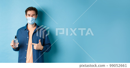Coronavirus, quarantine and social distancing concept. Happy young guy in medical mask recommending bottle of hand sanitizer, pointing at antiseptic and smiling, blue background Coronavirus, quarantine and social distancing concept. Happy young guy in medical mask recommending bottle of hand sanitizer, pointing at antiseptic and smiling, blue background 99052661