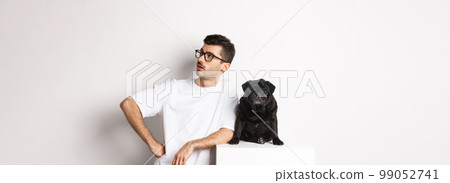 Handsome young man standing near cute black pug, looking right with arrogant expression, standing over white background Handsome young man standing near cute black pug, looking right with arrogant expression, standing over white background 99052741