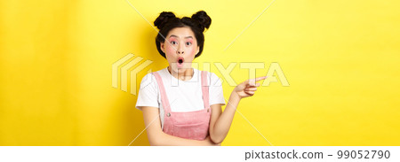 Surprised asian teen girl looking impressed, say wow and pointing finger left, standing with glamour makeup and summer clothes on yellow background Surprised asian teen girl looking impressed, say wow and pointing finger left, standing with glamour makeup and summer clothes on yellow background 99052790