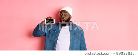 Online shopping and technology concept. Impressed african-american man in beanie showing phone screen, staring at mobile amazed, standing over pink background 99052833