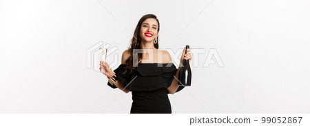 Celebration and party concept. Stylish brunette woman in glamour dress holding bottle and glass of champagne, smiling pleased, standing over white background 99052867