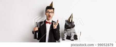 Happy young man celebrating holiday with cute dog, holding champagne and smiling, pug and owner wearing party costumes, white background 99052879