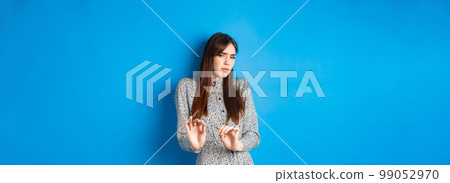 Disgusted and reluctant woman asking to keep away from here, showing block gesture, raising hands to stop you, standing on blue background 99052970