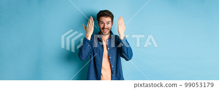 Happy young man open eyes and look surprised at gift, standing on blue background Happy young man open eyes and look surprised at gift, standing on blue background 99053179