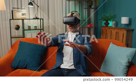 Old man using virtual reality futuristic technology headset to play simulation 3D video game at home 99054249