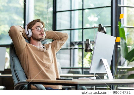 Young successful man in office resting with eyes closed and hands behind head, programmer finished work satisfied with achievement results and listening to music in headphones and podcast online. Young successful man in office resting with eyes closed and hands behind head, programmer finished work satisfied with achievement results and listening to music in headphones and podcast online. 99054721