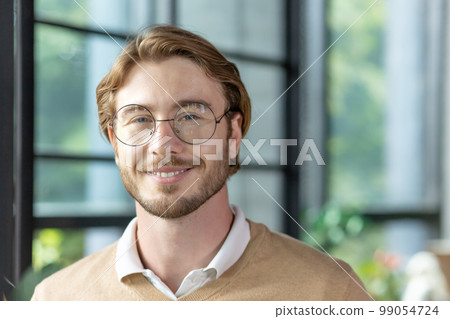 Close-up portrait of a young man inside the office, a businessman in glasses is smiling and looking at the camera, a blond programmer in casual clothes with a beard. 99054724