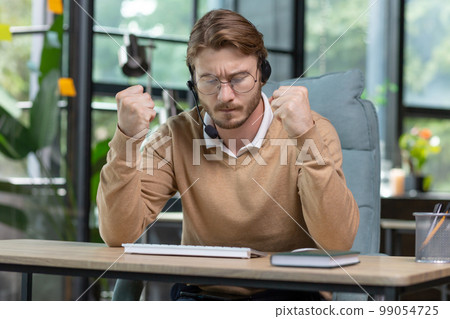 Angry office worker working inside office, young businessman in casual clothes with headset for video call, male nervous tech support worker and customer consultation online. 99054725