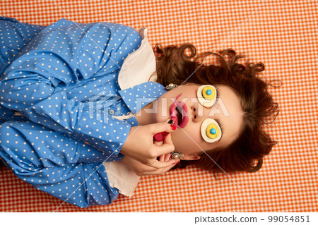 Doing makeup. Weird people concept. Young girl lying on table with eggs on her eyes over peach color background. Vintage, retro style. Food pop art photography. 99054851