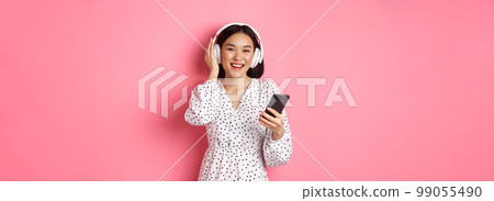 Beautiful asian woman listening music in headphones, using mobile phone, smiling happy at camera, standing over pink background 99055490