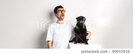 Amazed young man in glasses hugging his dog, pet owner and pug staring at upper left corner promo offer, standing over white background 99055638