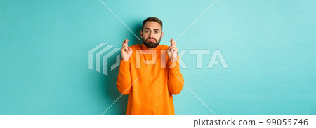 Hopeful sad man making a wish, cross fingers for good luck and sulking, standing over turquoise background Hopeful sad man making a wish, cross fingers for good luck and sulking, standing over turquoise background 99055746
