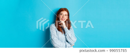 Thoughtful teenage girl with red hair, looking upper right corner logo and thinking, imaging something, standing over blue background 99055785
