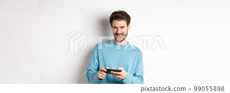 Image of handsome young man playing mobile video game, holding smartphone horizontally and smiling at camera pleased, standing over white background Image of handsome young man playing mobile video game, holding smartphone horizontally and smiling at camera pleased, standing over white background 99055898