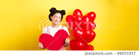 Happy Valentines day. Silly and beautiful asian woman smiling dreamy, showing red heart, imaging romantic date with lover, standing on yellow background Happy Valentines day. Silly and beautiful asian woman smiling dreamy, showing red heart, imaging romantic date with lover, standing on yellow background 99055937