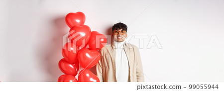 Valentines day. Excited young man smiling, looking hopeful, standing near big red hearts balloons, waiting for true love on lovers date, white background 99055944
