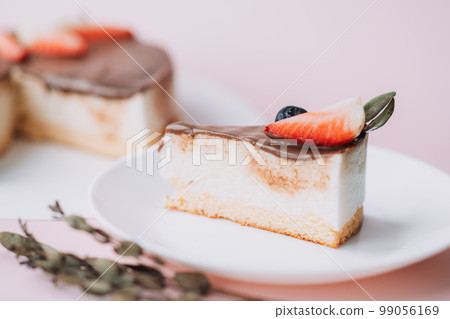 Bird's milk cake with chocolate decorated with strawberries and grapes with piece on white plate on pink background Bird's milk cake with chocolate decorated with strawberries and grapes with piece on white plate on pink background 99056169
