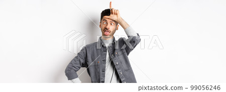 Funny young man making faces and showing tongue with loser sign on forehead, squinting eyes, standing on white background Funny young man making faces and showing tongue with loser sign on forehead, squinting eyes, standing on white background 99056246