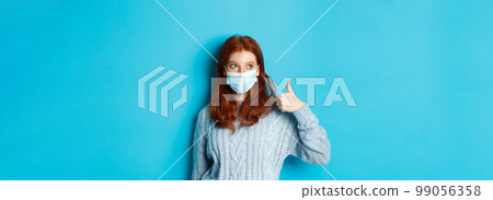Winter, covid-19 and social distancing concept. Satisfied young redhead woman in face mask showing alright, thumb up gesture and looking left at promo, blue background Winter, covid-19 and social distancing concept. Satisfied young redhead woman in face mask showing alright, thumb up gesture and looking left at promo, blue background 99056358