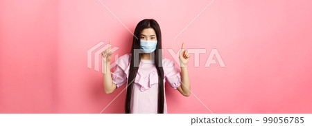 Coronavirus, quarantine and lifestyle concept. Beautiful asian woman in sterile medical mask pointing fingers up, smiling with eyes and look confident, standing against pink background Coronavirus, quarantine and lifestyle concept. Beautiful asian woman in sterile medical mask pointing fingers up, smiling with eyes and look confident, standing against pink background 99056785