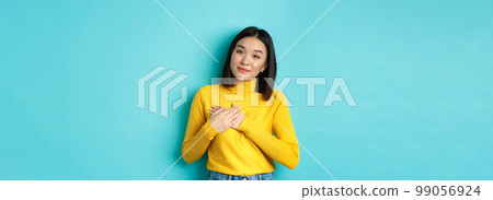 Portrait of beautiful heartfelt woman holding hands on heart, smiling and listening compassionate, standing over blue background in yellow pullover Portrait of beautiful heartfelt woman holding hands on heart, smiling and listening compassionate, standing over blue background in yellow pullover 99056924