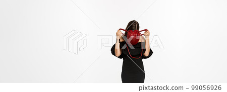Beauty and fashion concept. Full length of young woman sticking head inside purse and searching something, wearing black dress and high heels, standing over white background 99056926