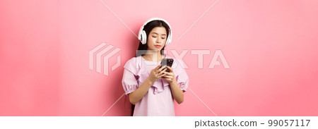 Young girl listening music in headphones and chatting on mobile phone, standing in dress against pink background Young girl listening music in headphones and chatting on mobile phone, standing in dress against pink background 99057117
