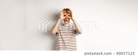 Impressed redhead man checking out promo offer, looking at camera from hand binoculars, smiling amazed, white background 99057125
