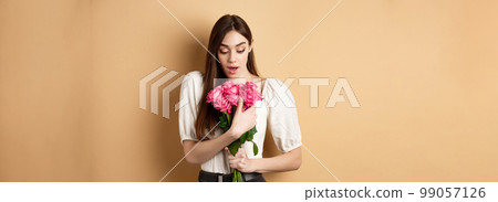 Valentines day. Surprised girl receive gift from lover on date, looking amazed at beautiful bouquet of flowers, holding roses on beige background Valentines day. Surprised girl receive gift from lover on date, looking amazed at beautiful bouquet of flowers, holding roses on beige background 99057126