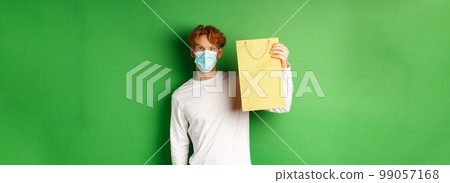 Covid-19 and pandemic concept. Surprised man in face mask showing shopping bag and looking at camera, standing over green background Covid-19 and pandemic concept. Surprised man in face mask showing shopping bag and looking at camera, standing over green background 99057168