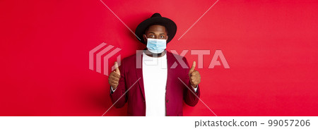 Covid-19 and fashion concept. Stylish african american man in hat and blazer, wearing face mask and showing thumb up, standing over red background 99057206