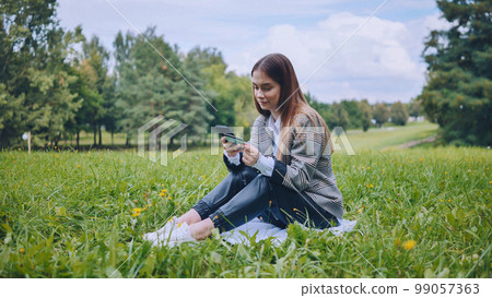A young girl makes a purchase with her phone and bank card in the park, sitting on the grass. A young girl makes a purchase with her phone and bank card in the park, sitting on the grass. 99057363