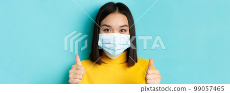 Covid-19, social distancing and pandemic concept. Close up of young asian woman in medical mask showing thumbs up, say yes, praise good offer, standing over blue background 99057465