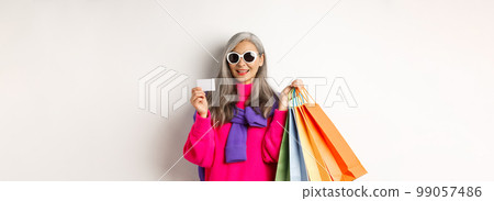 Stylish senior asian woman in sunglasses going shopping on holiday sale, holding paper bags and plastic credit card, standing over white background 99057486