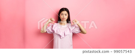 Skeptical and disappointed asian woman showing thumbs down with reluctant smirk, dislike product, show negative emotion, standing on pink background Skeptical and disappointed asian woman showing thumbs down with reluctant smirk, dislike product, show negative emotion, standing on pink background 99057543